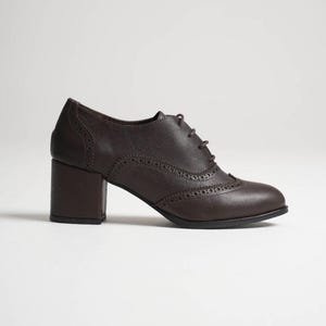 May include: A dark brown leather oxford shoe with a block heel. The shoe features decorative perforations along the seams and a lace-up closure. The heel measures approximately 2.5 inches.