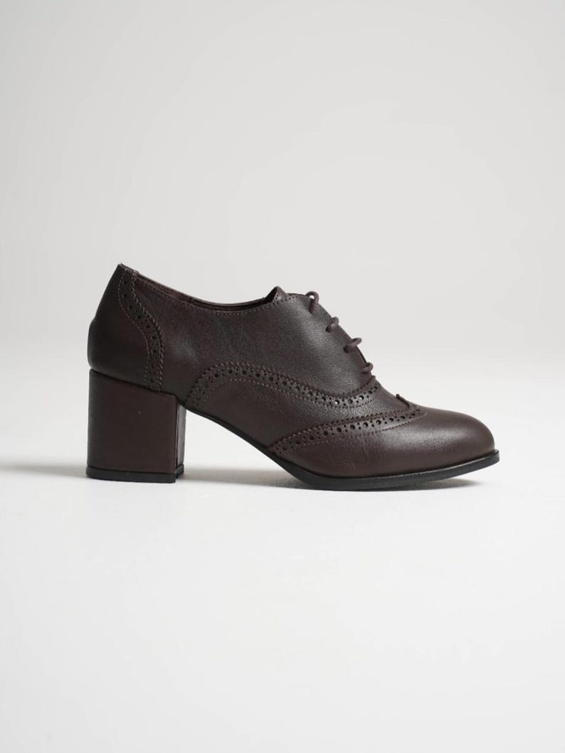May include: A dark brown leather oxford shoe with a block heel. The shoe features decorative perforations along the seams and a lace-up closure. The heel measures approximately 2.5 inches.