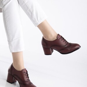 May include: Burgundy leather oxford-style shoes with a block heel. The shoes feature decorative perforations and lace-up closures. The heels are approximately 3 inches high. The shoes are worn with white pants.