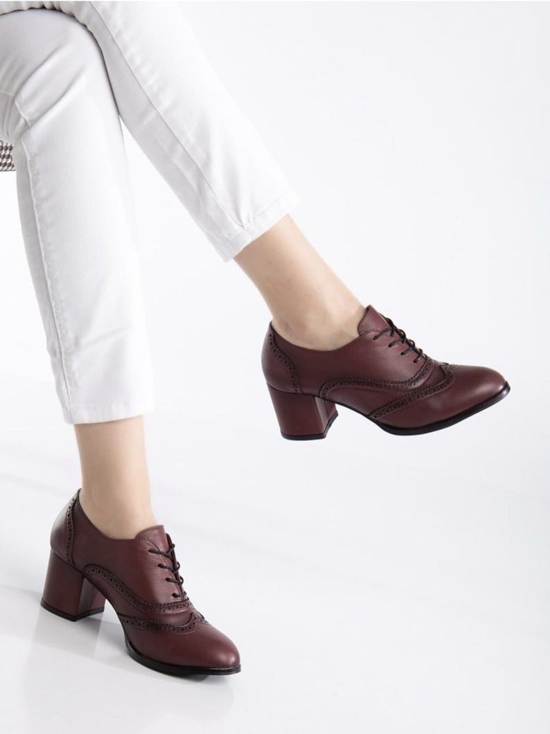 May include: Burgundy leather oxford-style shoes with a block heel. The shoes feature decorative perforations and lace-up closures. The heels are approximately 3 inches high. The shoes are worn with white pants.
