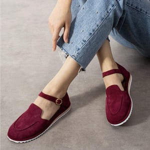 May include: A pair of deep burgundy suede loafers featuring a rounded toe, a thin ankle strap with a small gold-toned buckle, and a white serrated rubber sole. The shoes have a low-cut side profile and a stitched seam detail across the vamp.