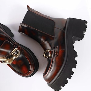 May include: A pair of brown and black ankle boots with a chunky platform sole. The boots feature a reddish-brown, glossy finish with gold-colored hardware and a black elastic panel. The boots are a stylish footwear option.