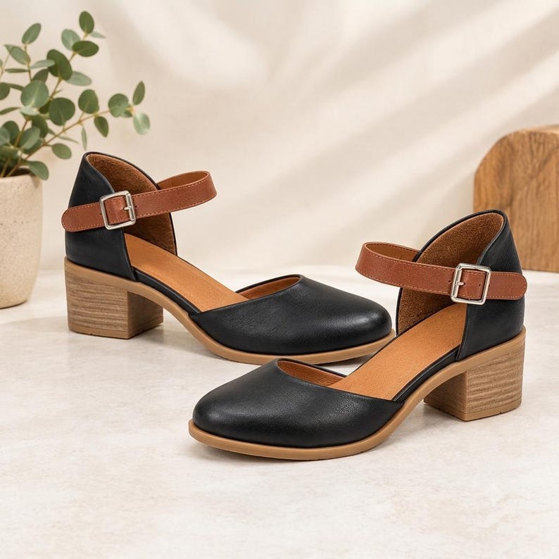 May include: A pair of black shoes with a rounded toe, featuring a tan ankle strap with a silver-toned metal buckle and a light brown block heel. The interior of the shoes is tan, and the outer material has a smooth, matte finish.
