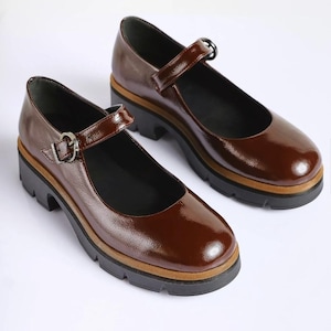May include: A pair of dark brown patent leather Mary Jane shoes featuring a rounded toe, a single adjustable strap with a silver-toned metal buckle, a tan midsole, and a thick, black lugged rubber sole with a block heel for added height.