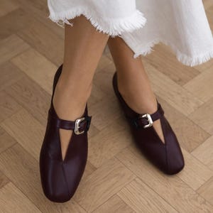 May include: Burgundy leather flats with a silver buckle closure. The shoes have a V-shaped cutout on the top and are worn with white linen pants with a fringed hem. The shoes are on a wooden floor.