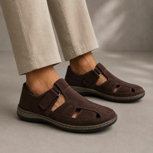 May include: A pair of dark brown suede closed-toe sandals featuring side cutouts, a hook-and-loop strap closure across the instep, and a dark grey rubber sole. The footwear is worn with light beige trousers that end above the ankle.