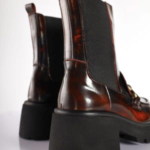 May include: A pair of brown leather ankle boots with a glossy finish. The boots have a black elastic panel on the side and a chunky black sole. The boots have a pull tab at the top and gold-colored hardware.