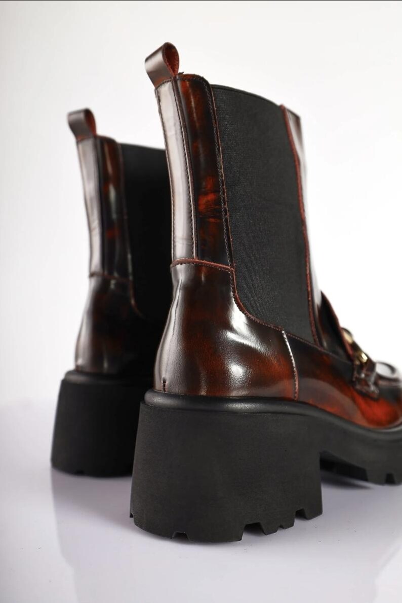May include: A pair of brown leather ankle boots with a glossy finish. The boots have a black elastic panel on the side and a chunky black sole. The boots have a pull tab at the top and gold-colored hardware.