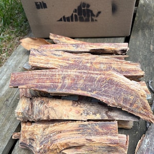 May include: A collection of brown, textured wooden pieces with reddish-brown streaks. The wood is arranged on a weathered wooden surface. A cardboard box with a state outline stencil is in the background.