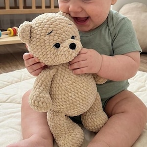 May include: A beige crochet teddy bear made from soft, chunky yarn. The stuffed animal features a rounded head with small ears, a dark brown stitched nose, and two black plastic safety eyes. The body has a textured, looped stitch pattern.