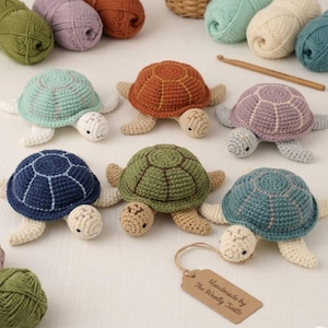 May include: Six crocheted turtles in various colors including mint, rust, lavender, navy, olive, and teal, each with a matching shell pattern. A wooden crochet hook and several yarn balls are nearby. A tag reads "Handmade by The Woolly Turtle."