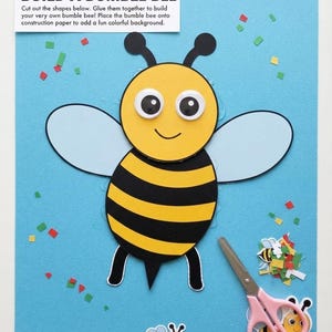 May include: A paper craft project titled BUILD A BUMBLE BEE on a light blue background. The bee features a yellow head with googly eyes, a black and yellow striped body, black antennae, and light blue wings. Pink-handled scissors are included.