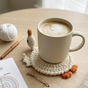 Peut inclure: Un dessous de verre crocheté en forme de cygne couleur crème avec un bec et des pattes orange, supportant une tasse en céramique remplie d'un latte. Le dessous de verre présente un corps circulaire, un cou et une tête. Une étiquette ronde THE SPIRAL SWAN est attachée par une ficelle.