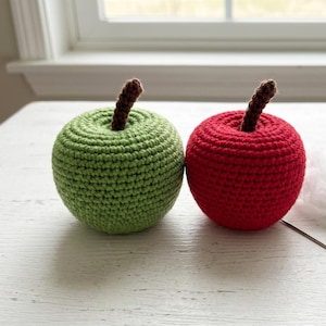 May include: Two crocheted apples, one in bright green and one in vibrant red, each featuring a small, textured brown stem attached to the top. The yarn work displays a consistent, tight stitch pattern throughout the rounded fruit shapes.