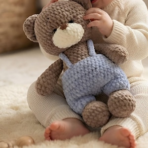 May include: A brown crocheted teddy bear toy with a cream-coloured muzzle and a small brown nose. The bear is wearing light blue crocheted overalls with shoulder straps. The plush toy has a soft, textured yarn finish and rounded ears.