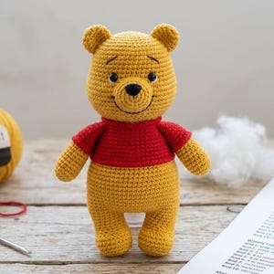 Puede incluir: Un oso de ganchillo con extremidades y cabeza de hilo amarillo, vistiendo una camiseta roja. El oso tiene ojos y nariz bordados en negro. A su lado hay un ovillo de hilo amarillo, un ovillo pequeño rojo, un ganchillo de metal y relleno blanco.