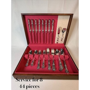 Puede incluir: Un cofre de madera con interior de terciopelo rojo, que contiene un juego de cubiertos de plata ornamentados de 44 piezas. El juego incluye cuchillos, tenedores y cucharas, dispuestos ordenadamente. El texto "Service for 8" y "44 pieces" es visible.