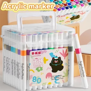 Puede incluir: Una caja de plástico transparente que contiene 80 rotuladores de pintura acrílica. Los rotuladores son de varios colores, con barriles blancos y puntas de colores. La caja tiene un asa y una etiqueta con texto y una ilustración de oso de dibujos animados. El texto "Acrylic marker" está en la parte superior.