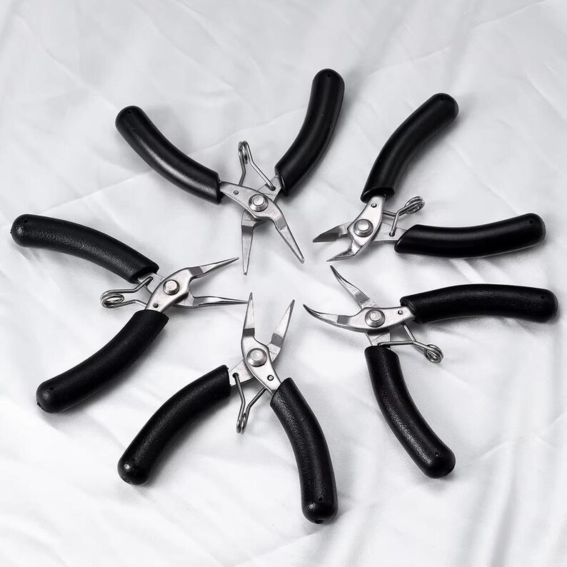 May include: A collection of six metal pliers with black, curved handles. The pliers are arranged in a circular pattern against a white background. The tools have a silver finish and are designed for precision work.