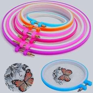 Könnte beinhalten: Eine Sammlung farbenfroher Stickrahmen in verschiedenen Größen, von Lila bis Blau, mit verstellbarer Metallhardware. Gezeigt werden auch zwei fertige Kreuzstichprojekte mit Schmetterlingsmotiven, eines in einem Rahmen und das andere separat.