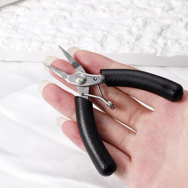 May include: A pair of silver-colored pliers with black rubber handles. The pliers have sharp, pointed tips and a spring mechanism. The tool is designed for precision work and is likely used for crafting or jewelry making.
