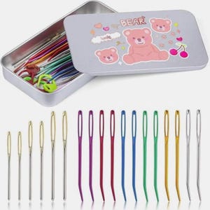 Op de afbeelding: Een set naaipatronen in verschillende maten en kleuren, gepresenteerd voor een zilveren blik. Het blik is voorzien van een roze berenillustratie en het woord "BEAR". De naalden zijn in een rij gerangschikt, sommige met gouden of zilveren oogjes.