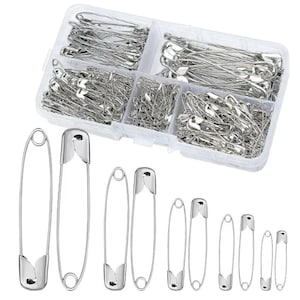 Könnte beinhalten: Ein klarer Kunststoff-Organizer gefüllt mit silbernen Sicherheitsnadeln in verschiedenen Größen. Mehrere größere Sicherheitsnadeln werden unterhalb des Organizers angezeigt. Diese Nadeln werden häufig zum Nähen, Basteln und für allgemeine Haushaltsaufgaben verwendet.