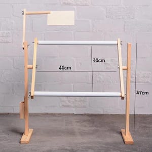 Puede incluir: Un soporte de bordado de madera con un marco blanco para punto de cruz. El soporte mide 47 cm de alto, con un marco de 40 cm de ancho y una altura de 30 cm entre los marcos. Tiene un pequeño letrero de madera en la parte superior.