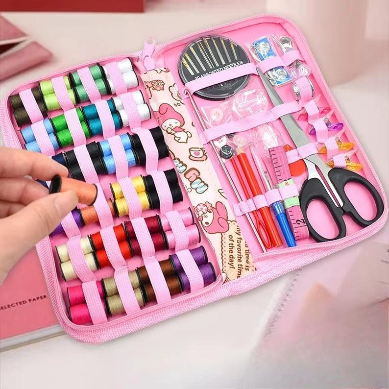 May include: A pink sewing kit with a variety of colorful thread spools, needles, scissors, and a measuring tape. The kit is open, revealing the organized contents. The kit includes a variety of sewing tools and accessories.