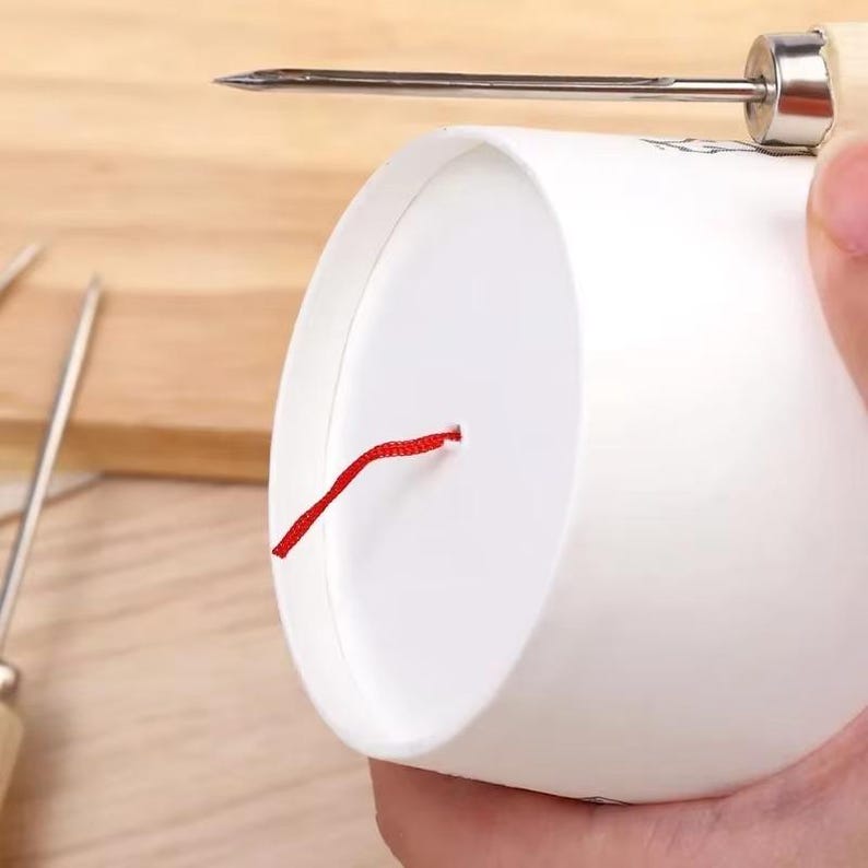 May include: A white paper cup with a red string emerging from a hole in the side. A metal awl with a wooden handle is positioned above the cup, with the sharp point directed towards the cup.