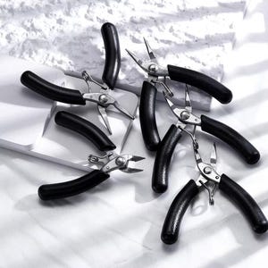May include: Several small, silver pliers with black handles are arranged on a white surface. The pliers have a curved handle design. These tools are likely for detailed work or crafts.