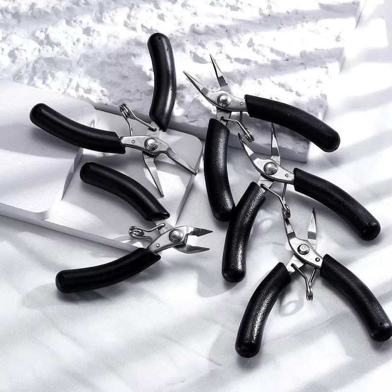 May include: A collection of small, silver-colored pliers with black handles. The pliers are scattered across a white surface, with some handles curved and others straight. The tools appear to be new.