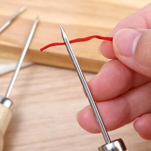 May include: Close-up of a silver needle with a wooden handle, piercing a strand of red thread. The needle is sharp and pointed, designed for sewing or crafting. The background is blurred, highlighting the needle and thread.