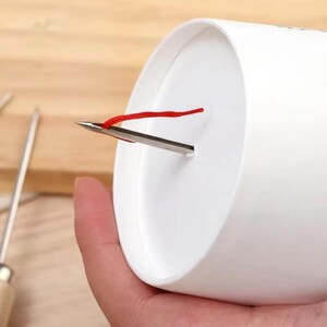 May include: A white cylindrical object with a metal needle and a red string attached. The needle is inserted through a small slit in the side of the cylinder. The background is a light brown wooden surface.