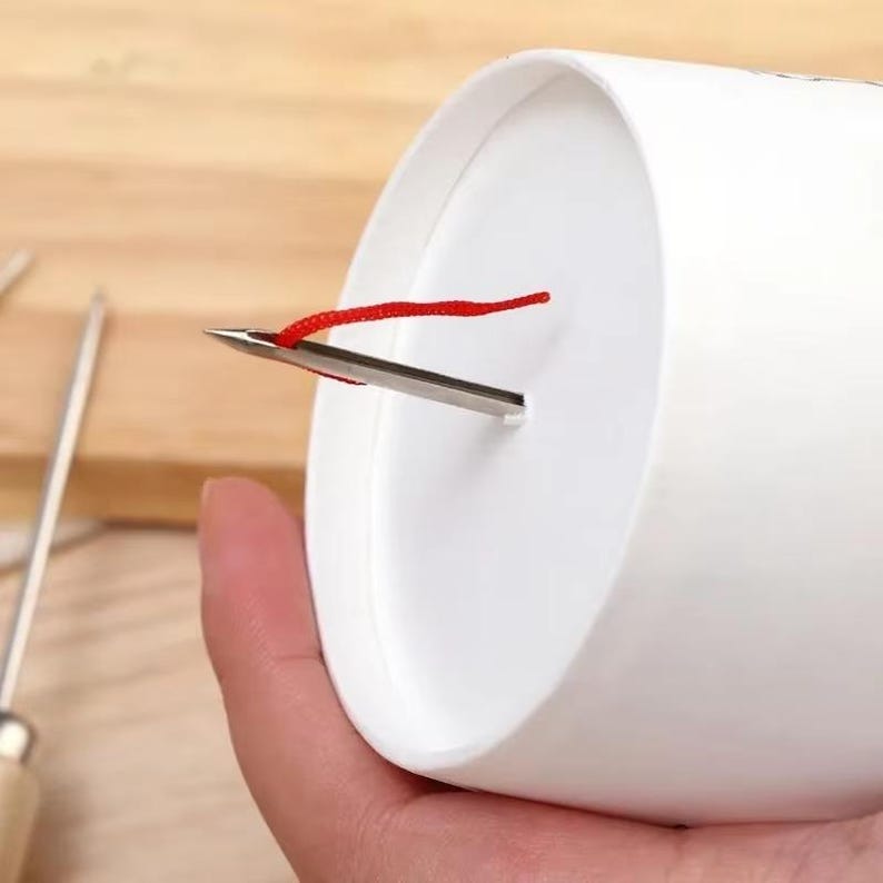 May include: A white cylindrical object with a metal needle and a red string attached. The needle is inserted through a small slit in the side of the cylinder. The background is a light brown wooden surface.