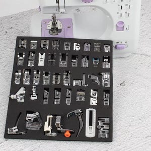 May include: A sewing machine with a variety of presser feet displayed in a black foam insert. The machine is white and purple. The presser feet are silver and come in various shapes and sizes, designed for different sewing tasks.