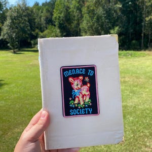 Può includere: Un libro color crema con angoli dorati. La copertina presenta una toppa con un cervo rosa, un fiocco blu e le parole "MENACE TO SOCIETY" in un font retrò. Il libro è tenuto all'aperto su uno sfondo di erba verde e alberi.