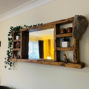 May include: A wooden mirror unit with integrated shelves, featuring small potted plants, decorative objects, and a plaid hat. The mirror reflects a room with a window and curtains. The wooden frame has a natural, unfinished appearance.