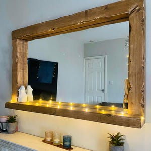 May include: A large rectangular mirror with a natural wood frame. The frame has a rough texture and a warm brown colour. The mirror is mounted on a white wall, with a shelf below adorned with fairy lights and decorative items.