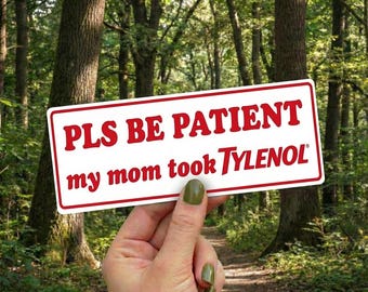 May include: A white rectangular sign with a red border and text that reads "PLS BE PATIENT my mom took TYLENOL" is held up in front of a forest background. The sign is held by a hand with green nail polish.