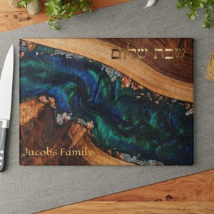 May include: Rectangular wooden cutting board with a river of blue and green resin. The board has the words "Shabbat Shalom" and "Jacobs Family" in gold lettering. The wood has a natural grain.
