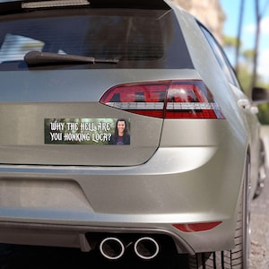 May include: A rectangular bumper sticker with the text "WHY THE HELL ARE YOU HONKING LOCA?" and a photo of a smiling person. The sticker is attached to the back of a silver car.