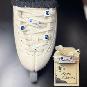 May include: Cream ice skate with white laces and blue jewelled accents. The skate has a silver charm that says "Salchow". A small wooden gift box with the words "Skate Charms" is also in the image.