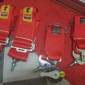 Puede incluir: Cuatro cinturones de seguridad de carreras rojos con hebillas de metal plateado y la marca "Sabelt" en etiquetas amarillas. Los cinturones est&aacute;n colocados sobre una superficie roja.