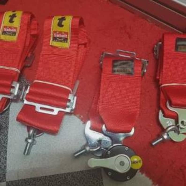 Puede incluir: Cuatro cinturones de seguridad de carreras rojos con hebillas de metal plateado y la marca "Sabelt" en etiquetas amarillas. Los cinturones est&aacute;n colocados sobre una superficie roja.