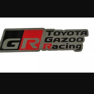 Könnte beinhalten: Ein metallisches Emblem mit dem Text "TOYOTA GAZOO Racing". Das Emblem hat eine rechteckige Form mit einem schwarzen und roten Design auf der linken Seite, mit den Buchstaben "GR" in Silber. Das Toyota Gazoo Racing Logo ist in Silber.