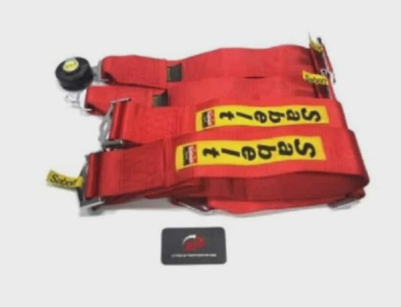 Puede incluir: Arn&eacute;s de carreras rojo con etiquetas amarillas y negras. El arn&eacute;s tiene hebillas met&aacute;licas y un bot&oacute;n de ajuste negro. Tambi&eacute;n hay una peque&ntilde;a tarjeta negra con un logotipo.