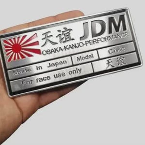 May include: A rectangular silver metal emblem with the text "JDM" and Japanese characters. The emblem features a red and white rising sun design and the text "OSAKA-KANJO-PERFORMANCE". It reads "Made in Japan" and "For race use only".