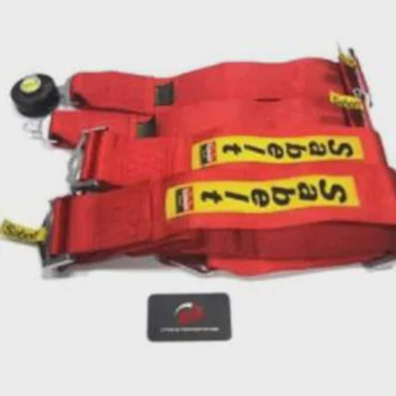 Puede incluir: Arn&eacute;s de carreras rojo brillante con texto "Sabelt" amarillo y negro. El arn&eacute;s presenta m&uacute;ltiples correas, hebillas met&aacute;licas y un bot&oacute;n de ajuste negro. Una peque&ntilde;a tarjeta negra con un logotipo est&aacute; en primer plano.