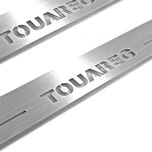 May include: Two brushed metal car door sill protectors with the word "TOUAREG" cut out. The metal has a brushed texture and a rectangular shape with a groove cut into the side. The background is white.
